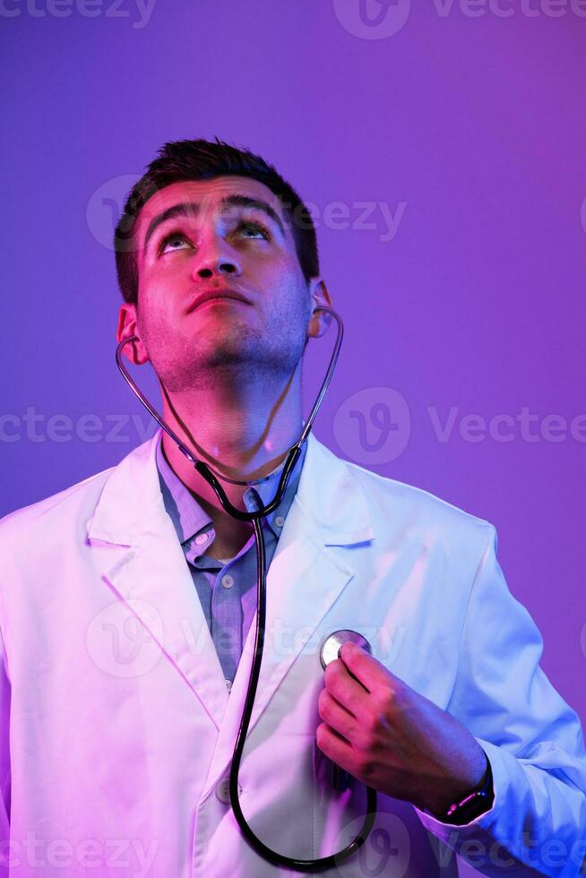 Porträt von Held im Weiß Mantel. heiter lächelnd jung Arzt mit Stethoskop im medizinisch Krankenhaus Stehen gegen Blau und Rosa Hintergrund. Coronavirus covid-19 Achtung warnen foto