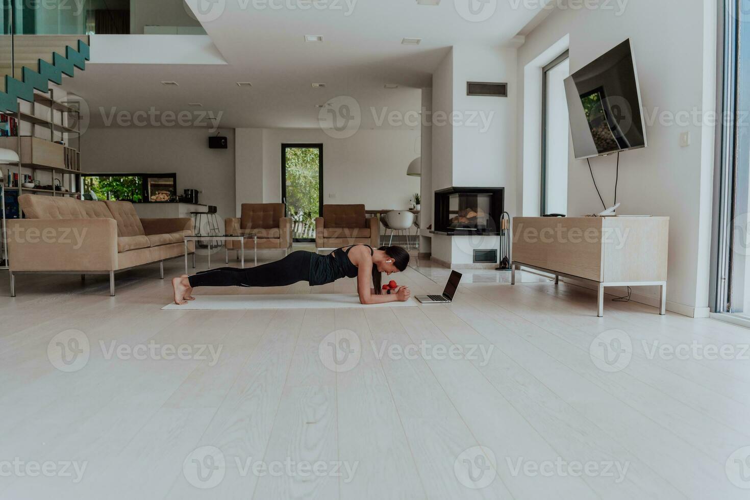 ein jung Frau im Sportbekleidung Arbeiten ein Planke im das Leben Zimmer während teilnehmen im online Ausbildung über ein Laptop foto