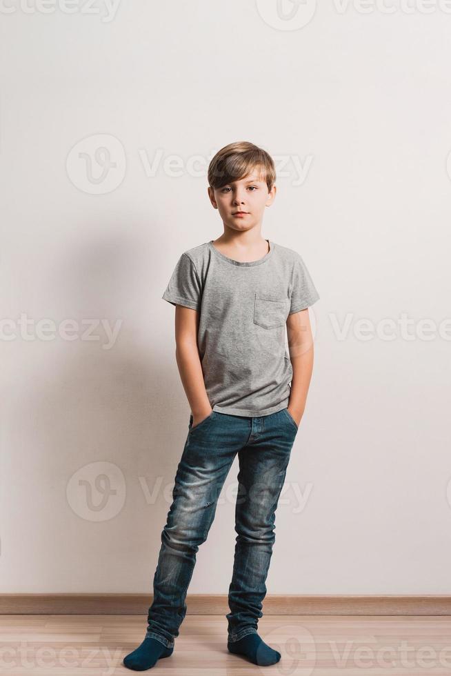 ein süßer Junge an der weißen Wand, graues Hemd, blaue Jeans in voller Größe foto