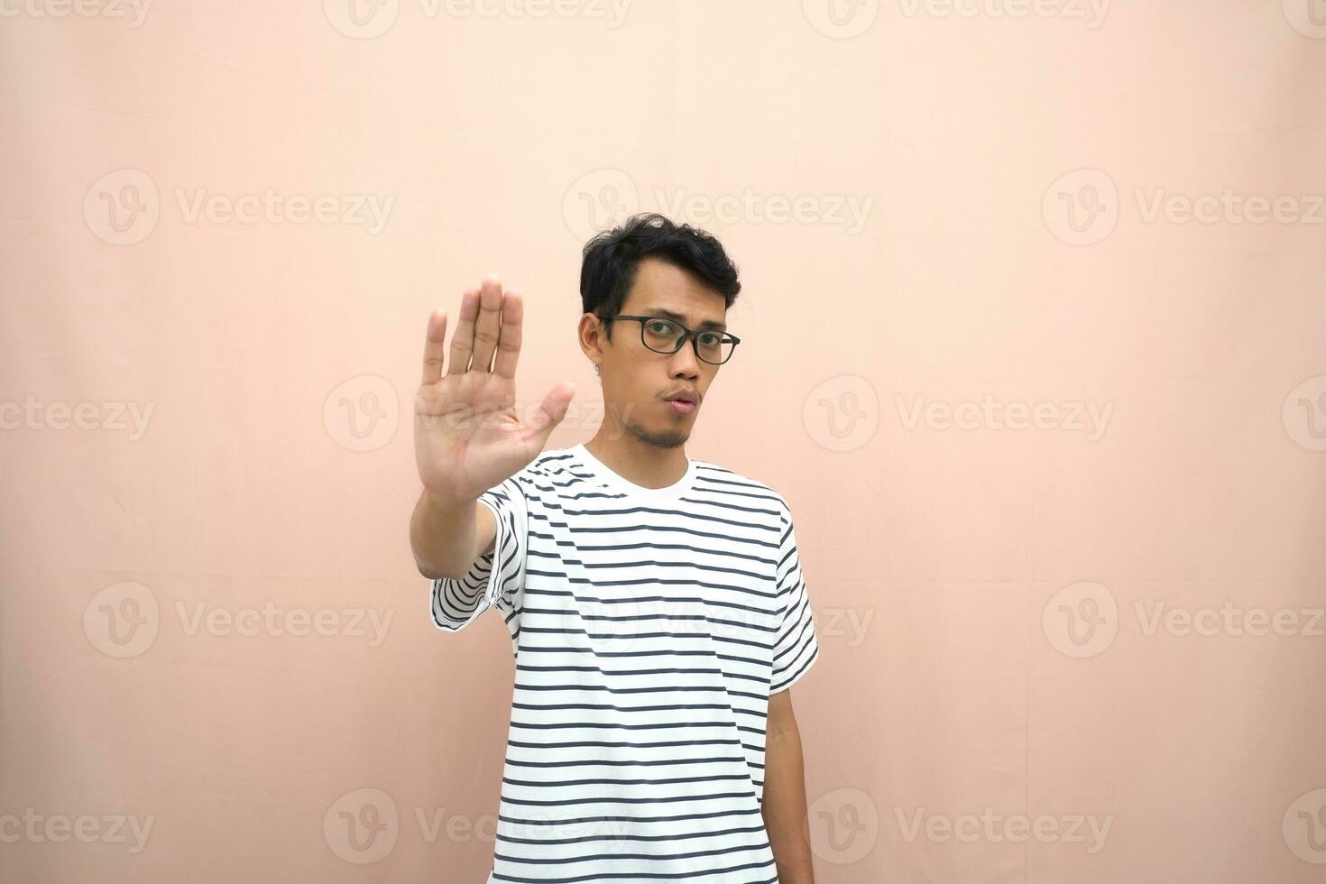 Porträt von ein asiatisch Mann im Brille tragen ein beiläufig gestreift T-Shirt. bezeichnen stoppen, Kreuz Verbot Zeichen, verbieten etwas, verweigern,. Beige Hintergrund. foto