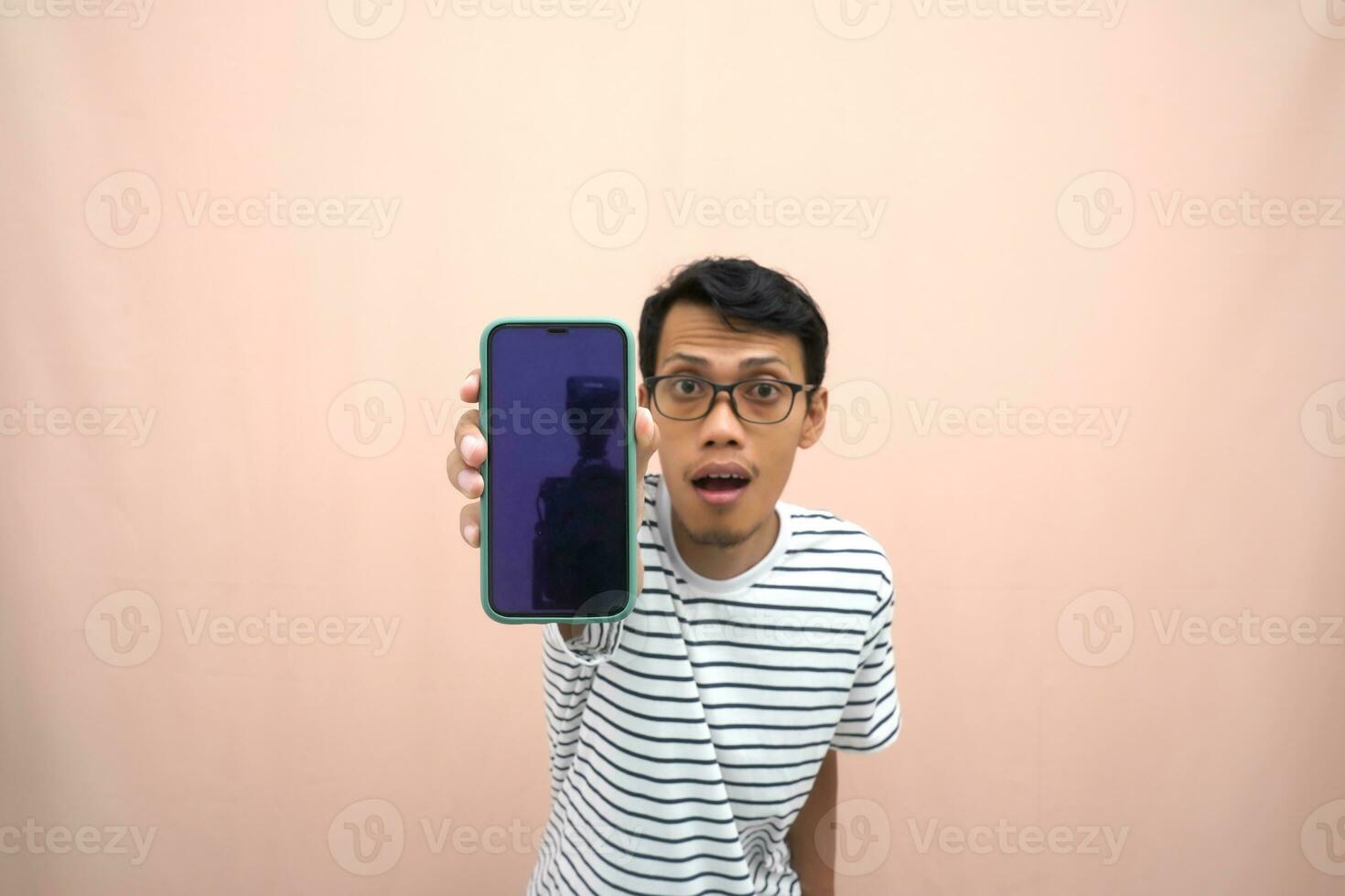 Porträt von ein asiatisch Mann tragen Brille tragen ein beiläufig gestreift T-Shirt. zeigt an Smartphone Bildschirm mit Glücklich, überrascht, glücklich Ausdruck, zum Förderung. isoliert Beige Hintergrund. foto