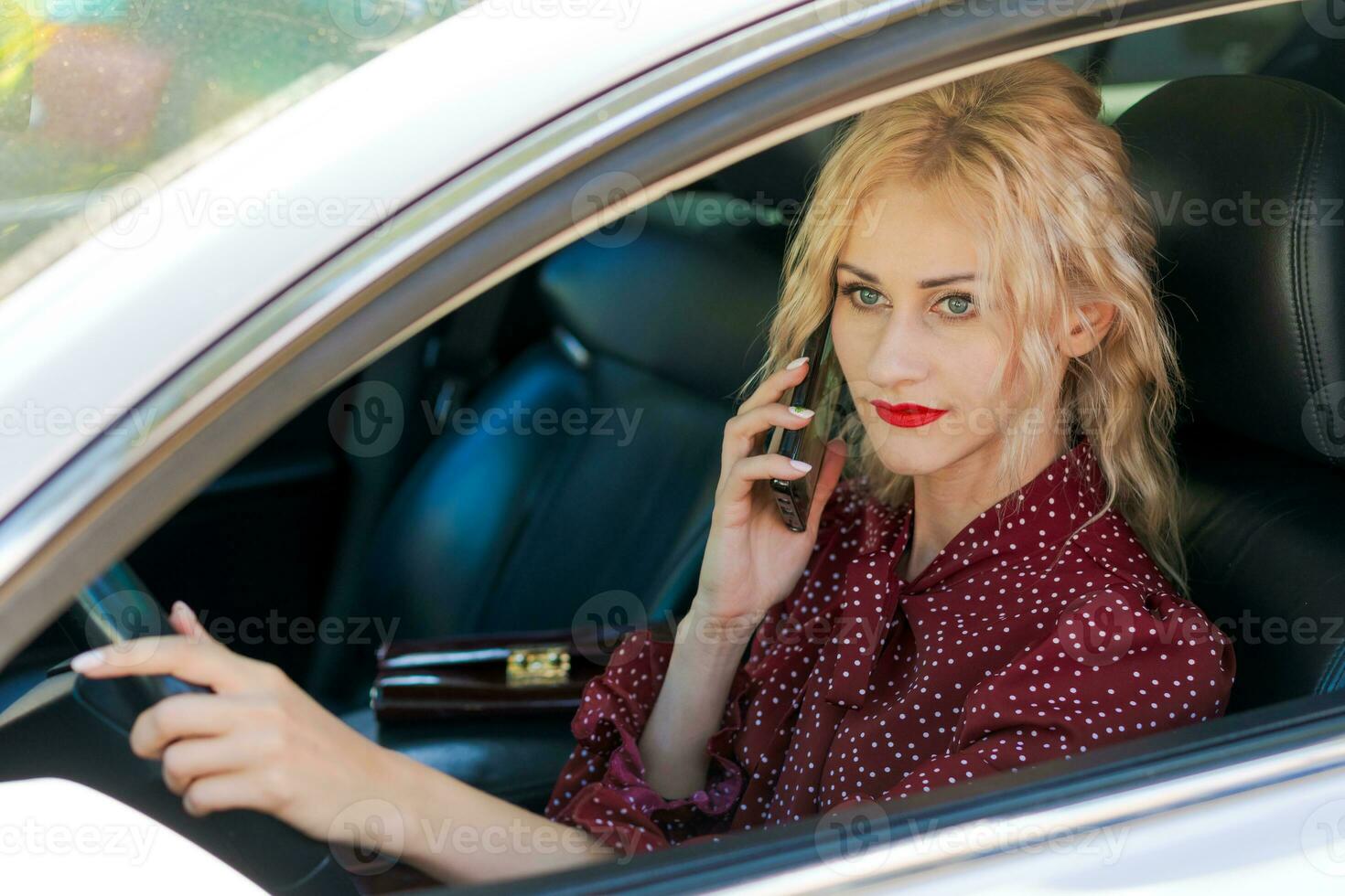 Porträt von ein schön blond Frau im ein rot Kleid Sitzung im ein Auto mit ein Telefon foto