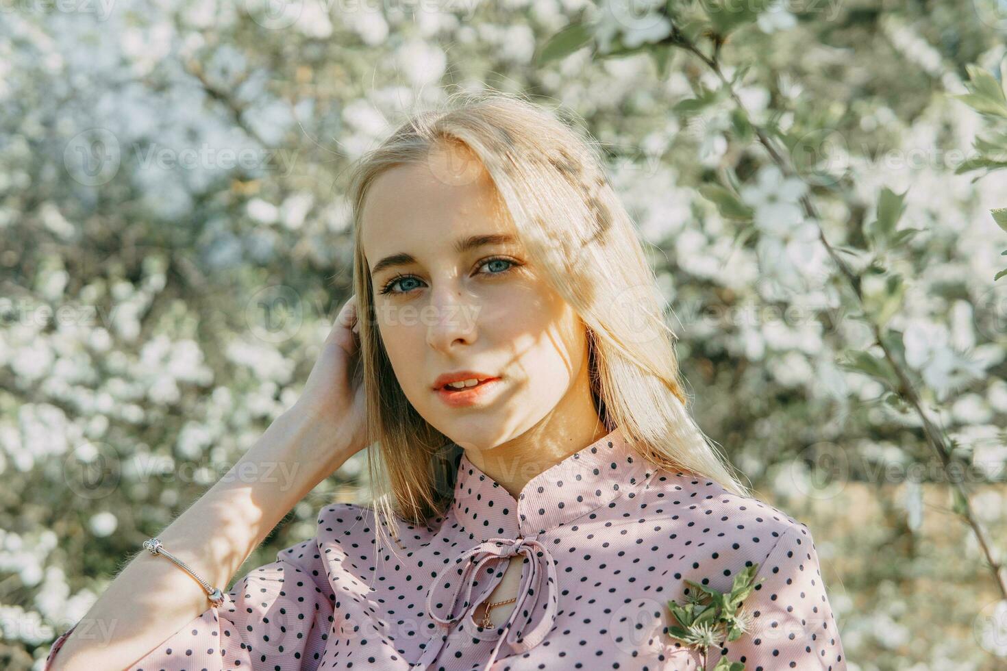 blond Mädchen auf ein Frühling gehen im das Garten mit Kirsche blüht. weiblich Porträt, Nahansicht. ein Mädchen im ein Rosa Polka Punkt Kleid. foto