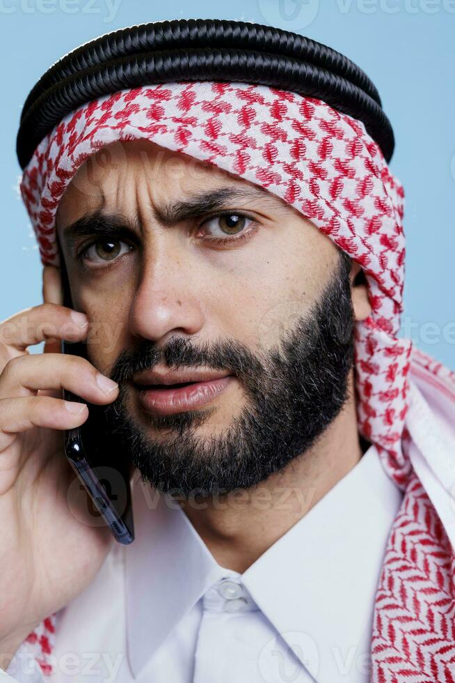 arabisch Mann runzelte die Stirn Augenbrauen und suchen beim Kamera mit verwirrt Ausdruck während chatten auf Smartphone. Muslim Person tragen ghutra Sprechen auf Handy, Mobiltelefon Telefon Nahansicht Porträt foto