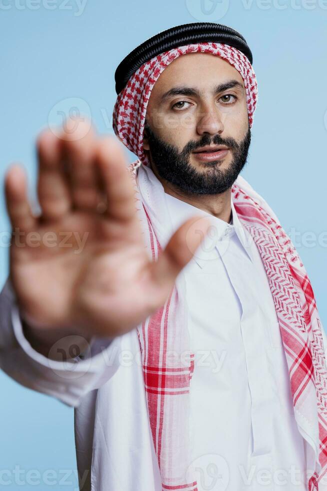 zuversichtlich Muslim Mann Herstellung verboten halt Zeichen mit Palme und suchen beim Kamera. ernst arabisch Person gekleidet im traditionell Kleider zeigen Ablehnung Geste mit Hand Studio Porträt foto