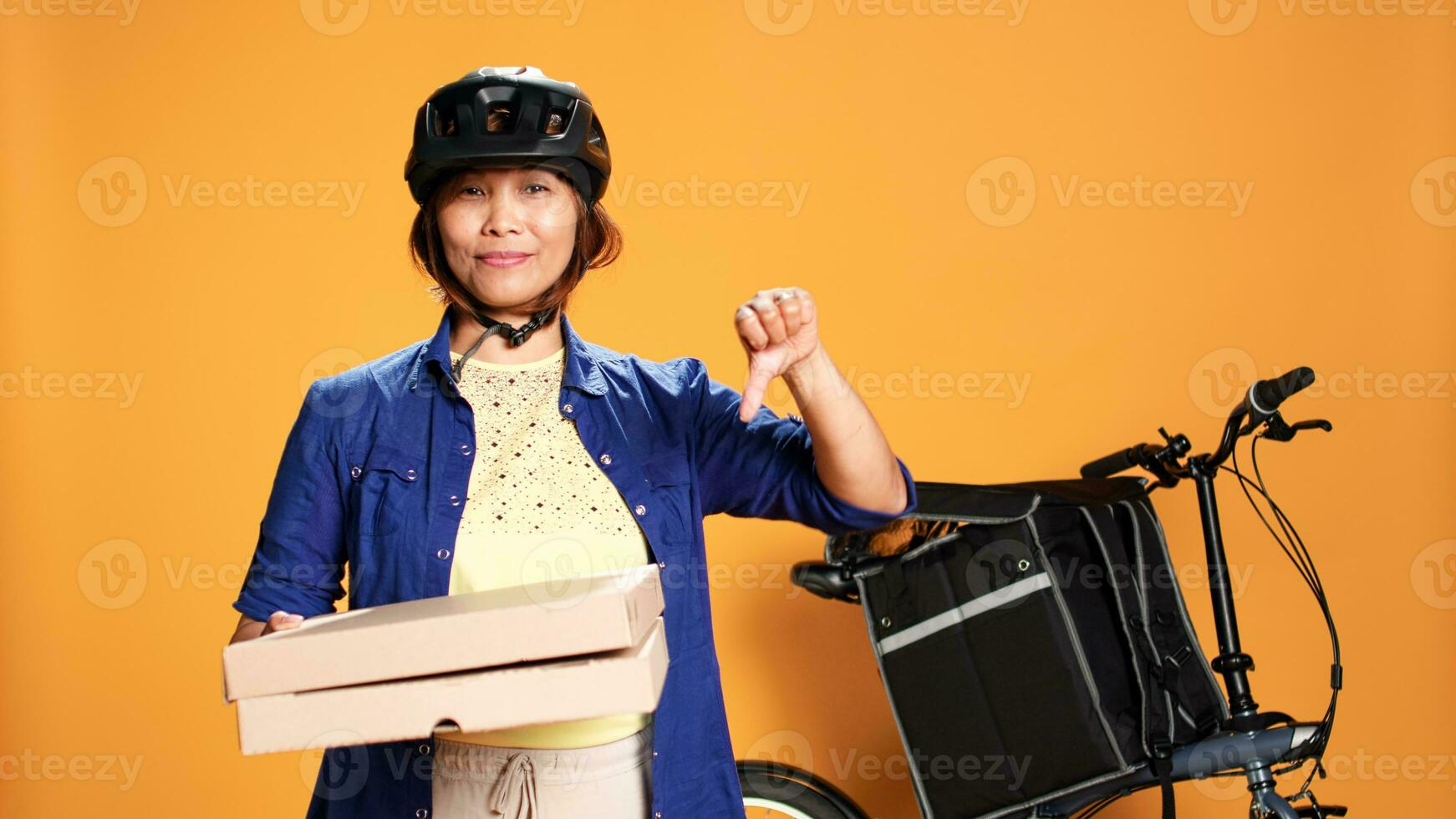 Porträt von Fachmann Kurier halten schnell Essen Pizza Kisten, tun missbilligend Aktion. Fahrrad Fahrer zeigen Daumen Nieder Zeichen während liefern wegbringen Mahlzeit, isoliert Über Studio Hintergrund foto