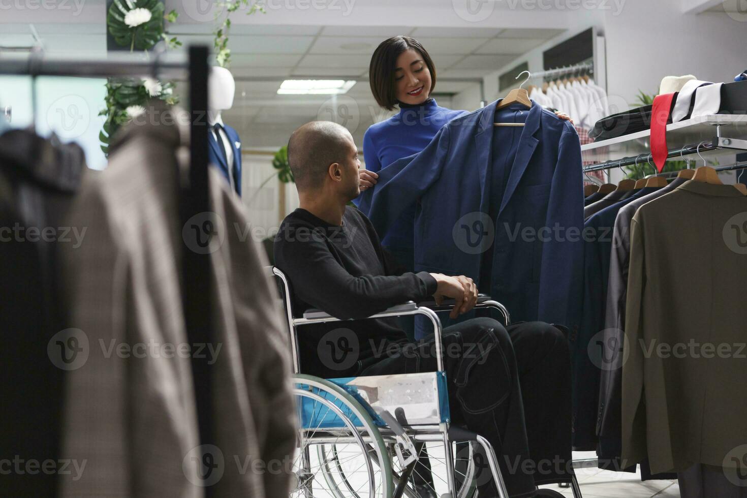 Kleidung Geschäft lächelnd Assistent Portion Kunde im Rollstuhl zu untersuchen Jacke Stil und fit. Einkaufen Einkaufszentrum asiatisch Frau Verkäufer Beratung arabisch Mann mit Behinderung und geben Mode Rat foto