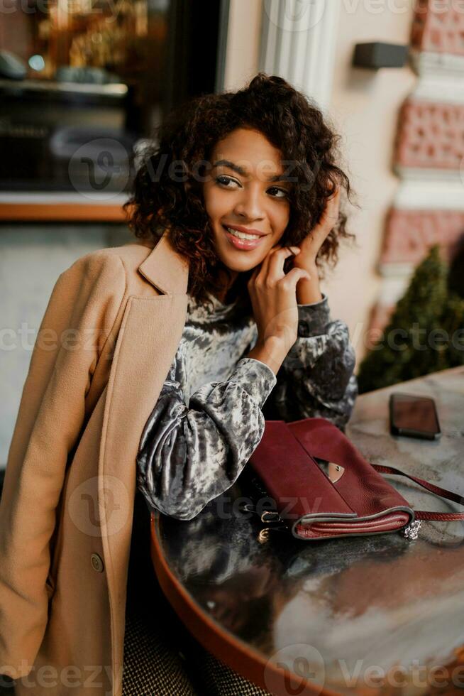 draussen Porträt von schön lächelnd schwarz Frau mit stilvoll afro Haare Sitzung im Cafe im Paris. tragen modisch Samt Kleid und Beige Mantel. Luxus Tasche. foto