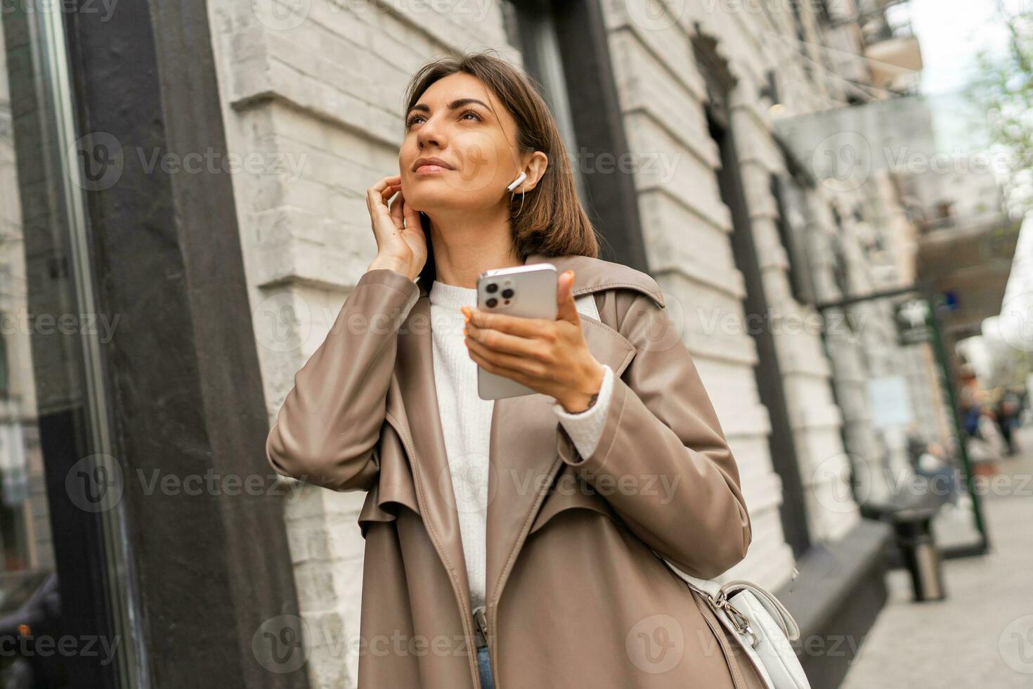 Straße Lebensstil Foto von stilvoll europäisch Geschäft Brünette Frau im Leder Mantel posieren draussen . modisch fallen Zubehör. mit Handy, Mobiltelefon Telefon.
