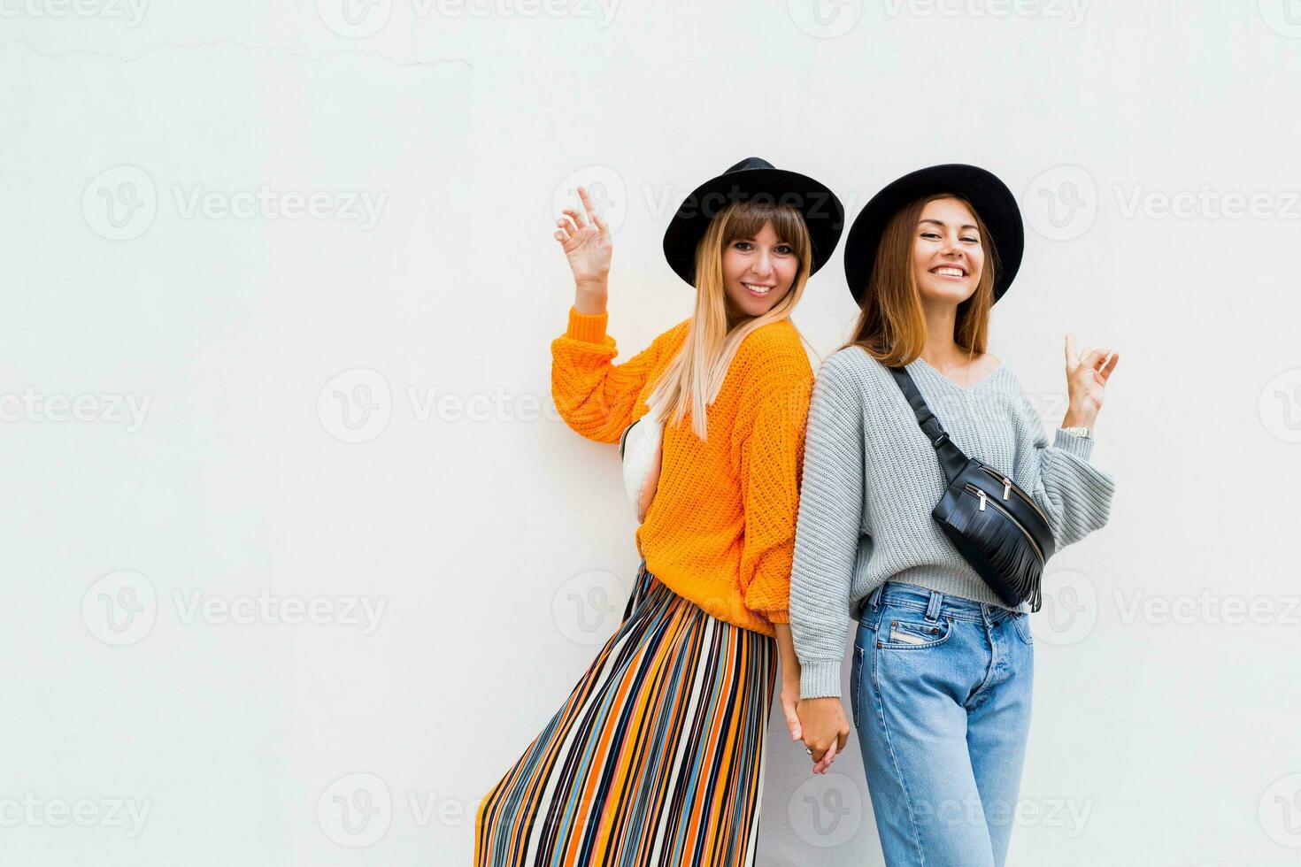 draussen Lebensstil Porträt von Paar von jung Frauen haben Spaß zusammen. windig Haare. zwei Mädchen täuschen um und Tanzen auf Weiß Hintergrund. stilvoll Gammler Taschen, ähnlich schwarz Hüte. foto