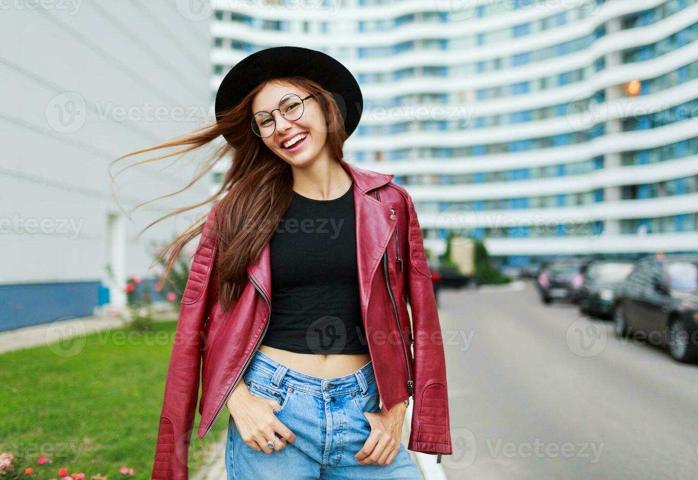 schön Mädchen mit offen Lächeln posieren auf das Straße . windig Haare . tragen runden Gläser, rot Leder Jacke. städtisch Hintergrund. foto