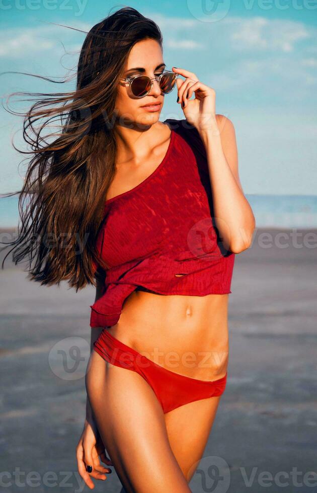 schön sexy schlank Frau im rot Bikini und T-Shirt mit Brille posieren auf das Strand gegen ein Blau Himmel. Sommer- Stimmung. foto