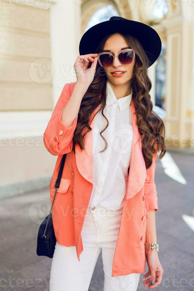draussen Mode Porträt von sexy stilvoll beiläufig Frau im schwarz Hut, Rosa passen , Weiß Bluse posieren auf alt Straße Hintergrund. Frühling , fallen sonnig Tag. wellig Frisur. foto