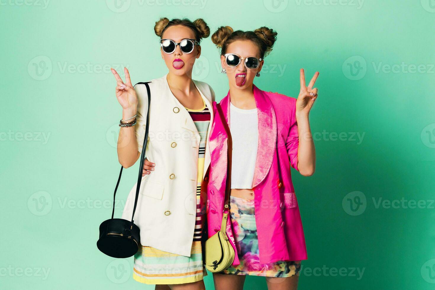 Mode Studio Bild von zwei jung Frauen im stilvoll beiläufig Frühling Outfit haben Spaß, Show Zunge. hell modisch Farben, stilvoll Frisur mit Gebäck , cool Sonnenbrille. freunde Porträt. foto