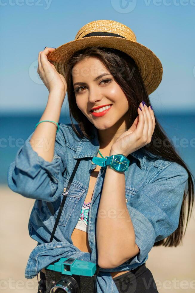 lächelnd Frau im Stroh Hut und stilvoll Sommer- Outfit posieren mit retro Kamera auf das Strand. foto