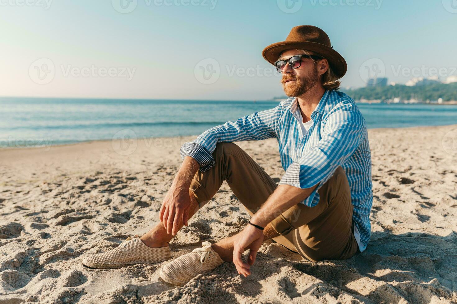 jung attraktiv Hipster Mann Sitzung auf Strand durch Meer auf Sommer- Ferien foto