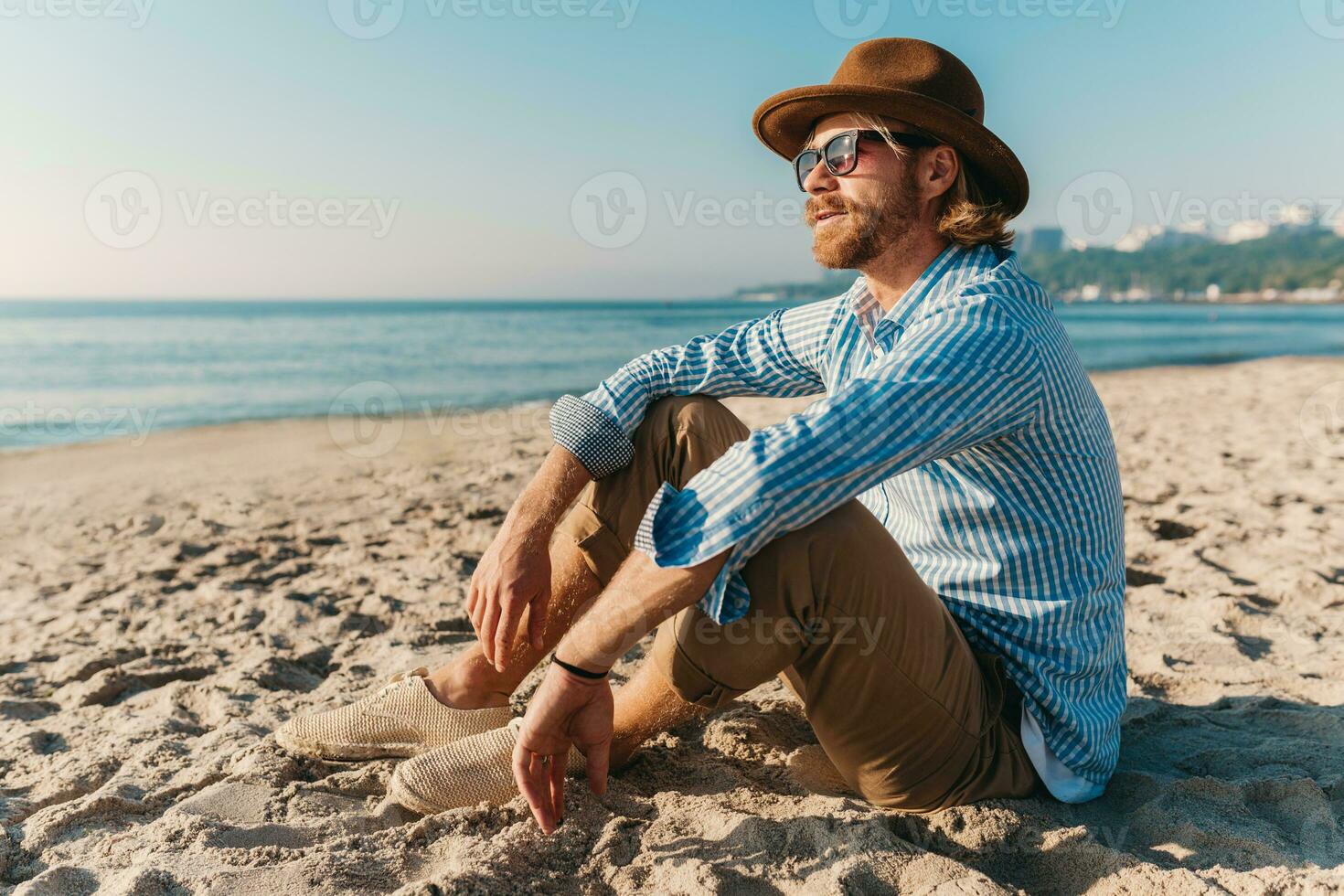 jung attraktiv Hipster Mann Sitzung auf Strand durch Meer auf Sommer- Ferien foto