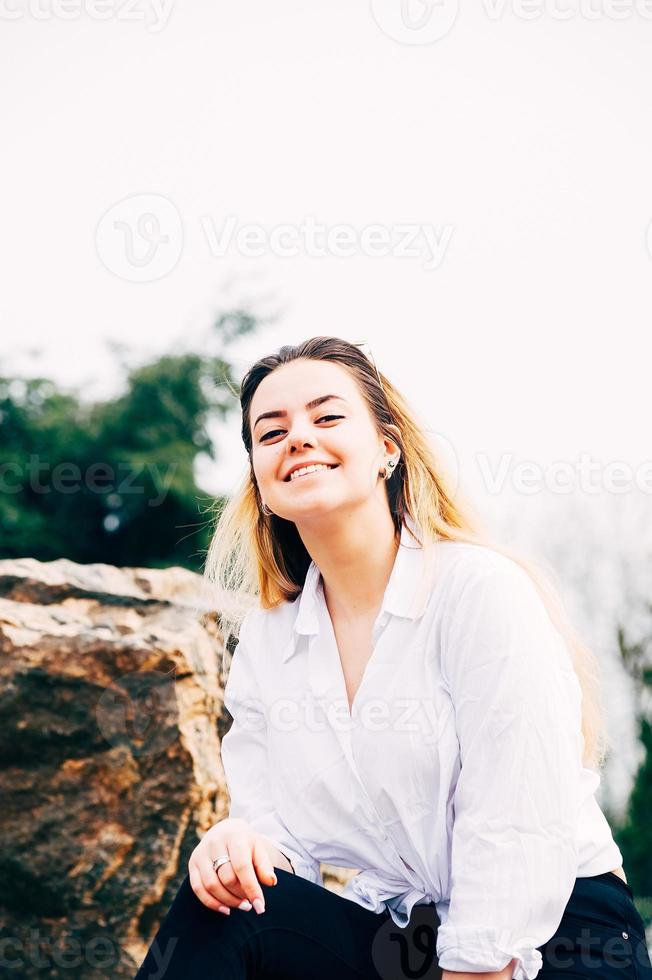 ein hübsches langhaariges Mädchen, das lächelt und auf einem großen Stein in einem Park sitzt foto