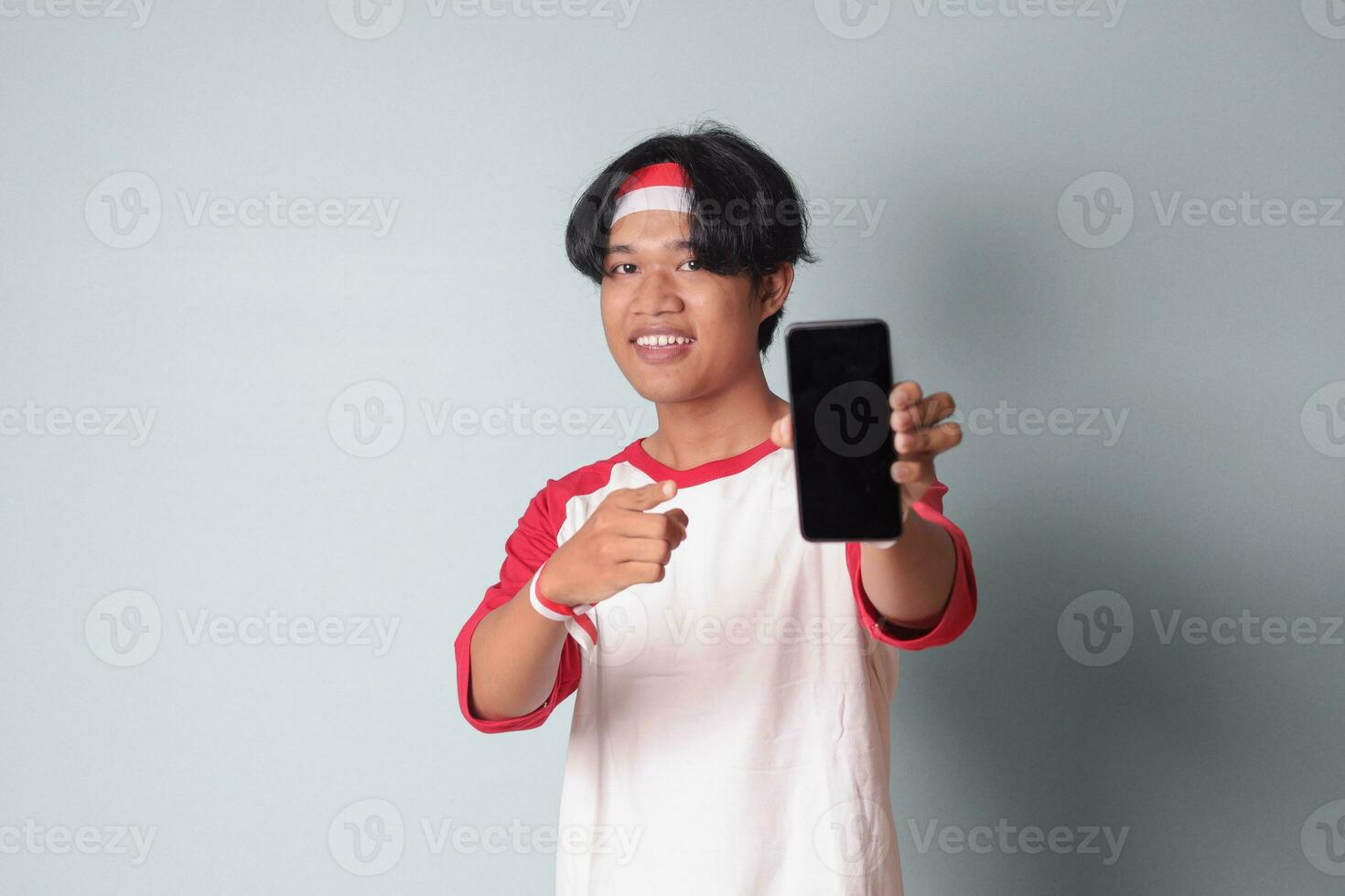 Porträt von attraktiv asiatisch Mann im T-Shirt mit rot und Weiß Band auf Kopf, halten und zeigen leer Bildschirm von Handy, Mobiltelefon Telefon zum Attrappe, Lehrmodell, Simulation. isoliert Bild auf grau Hintergrund foto