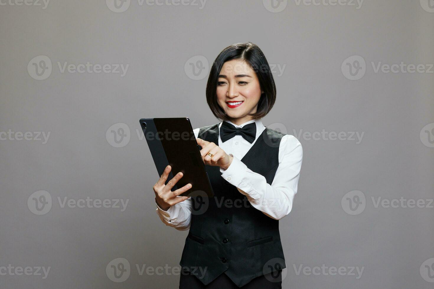 lächelnd Restaurant Rezeptionist im Uniform Überprüfung Gast aufführen auf Digital Tablette. Restaurant asiatisch Frau Kellnerin tippen auf Berührungssensitiver Bildschirm während Prüfung online bestellen während posieren im Studio foto