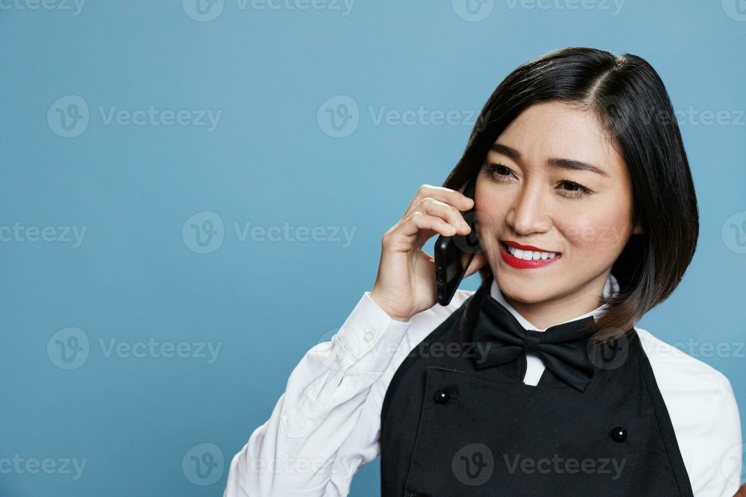 Hotel lächelnd heiter asiatisch Frau Rezeptionist tragen Uniform Sprechen auf Smartphone. froh Kellnerin antworten Anruf und chatten mit Gastronomie Bedienung Kunde auf Handy, Mobiltelefon Telefon foto