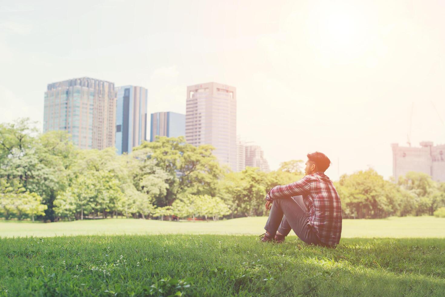 Rückansicht des entspannten jungen reisenden Mannes, der auf Gras im Park sitzt. foto