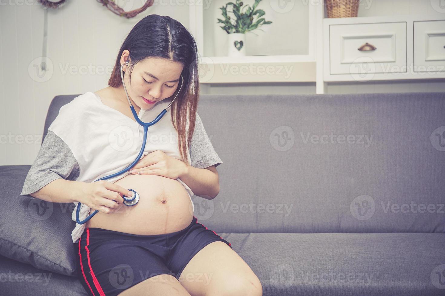 Nahaufnahme der schwangeren asiatischen Frau, die mit Stethoskop hört. foto