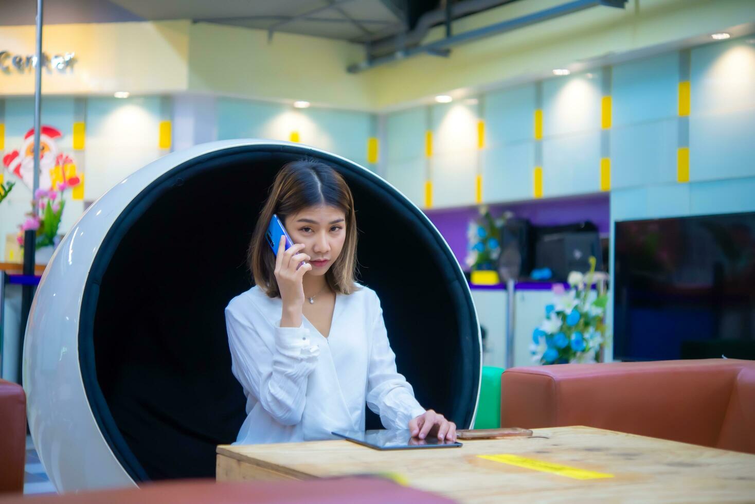 schön asiatisch Geschäft Frau Sitzung auf modern runden Stuhl und mit Telefon zu kommunizieren mit Kunde mit ernst Gesicht während suchen zu das Kamera, Digital Marketing. foto