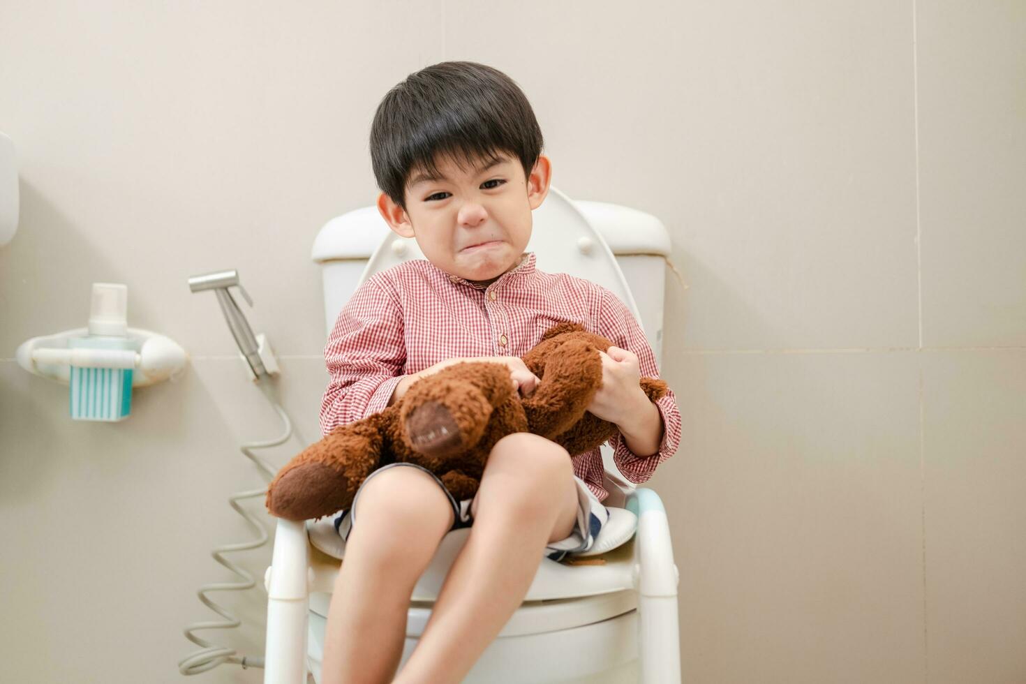 asiatisch Junge Sitzung auf das Toilette Schüssel im Hand halten Teddy Bär foto