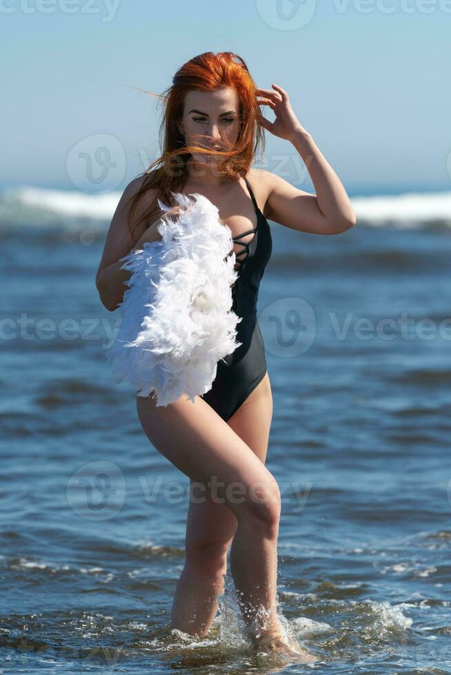 Rothaarige Frau auf Strand im Badeanzug Spaziergänge Knöchel tief in Wellen von Ozean, hält Feder Boa im Hand foto