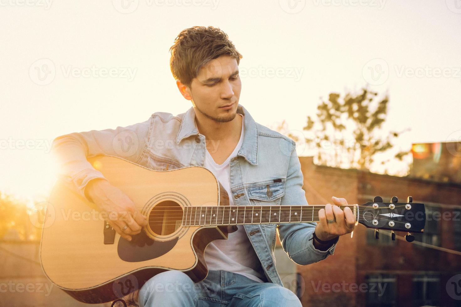 junger Mann, der Gitarre in der Stadt auf Sonnenstrahlenhintergrund spielt foto