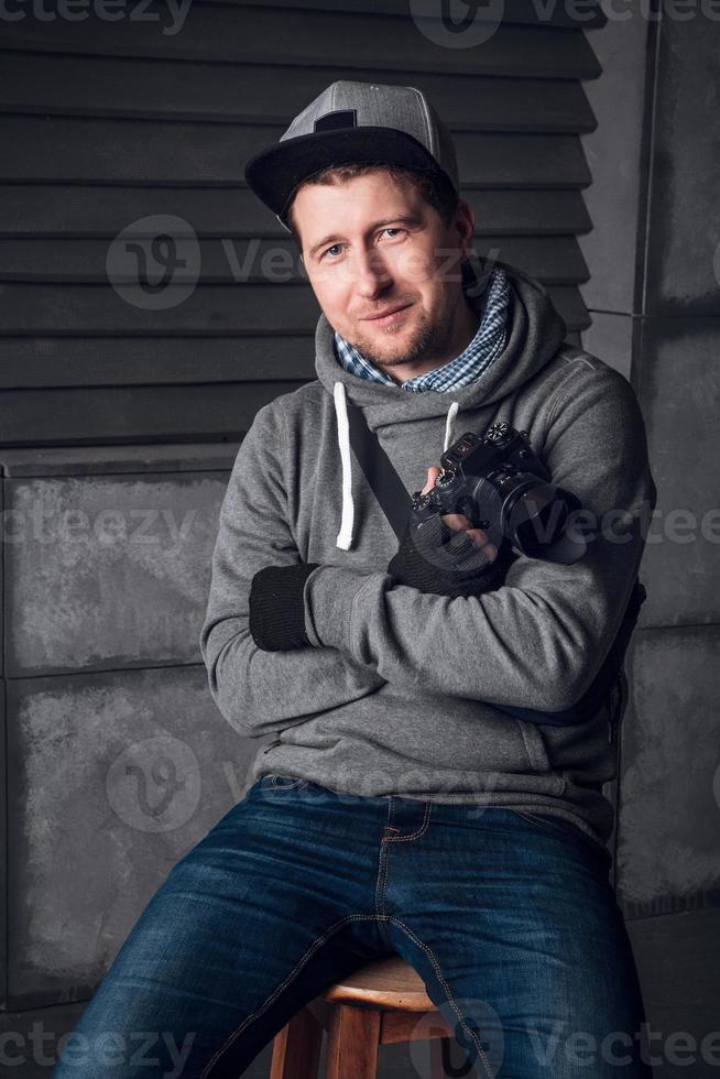 Mann mit Kamera und lustigem Gesicht sitzt auf einem Stuhl in einem grauen Studio foto