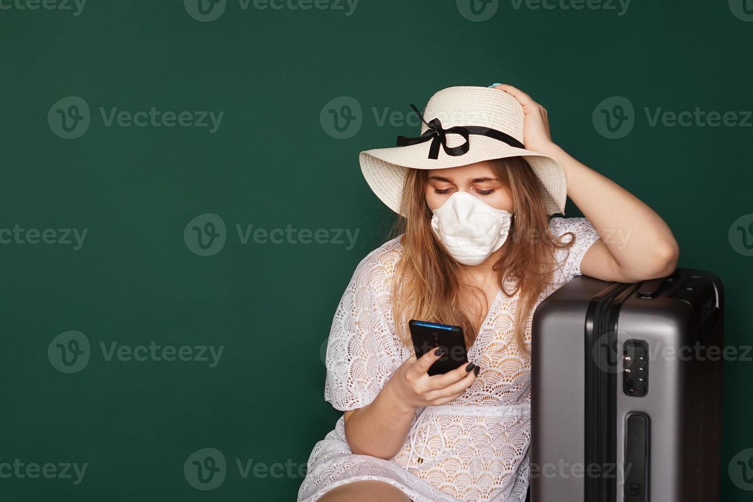 Mädchentourist in einer medizinischen Maske sitzt mit Gepäck auf grünem Hintergrund foto