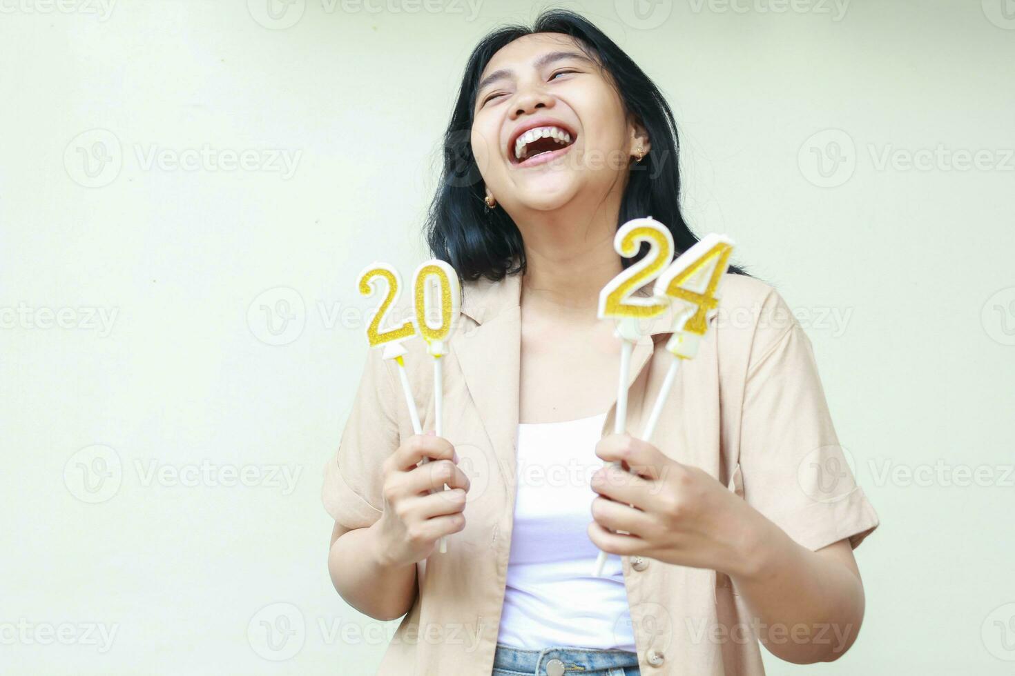 Porträt von glücklich asiatisch jung Frau Hipster Lachen mit öffnen Mund und suchen beiseite und oben während halten 2024 Zahl Kerzen tragen stilvoll braun äußere isoliert auf Weiß Hintergrund foto