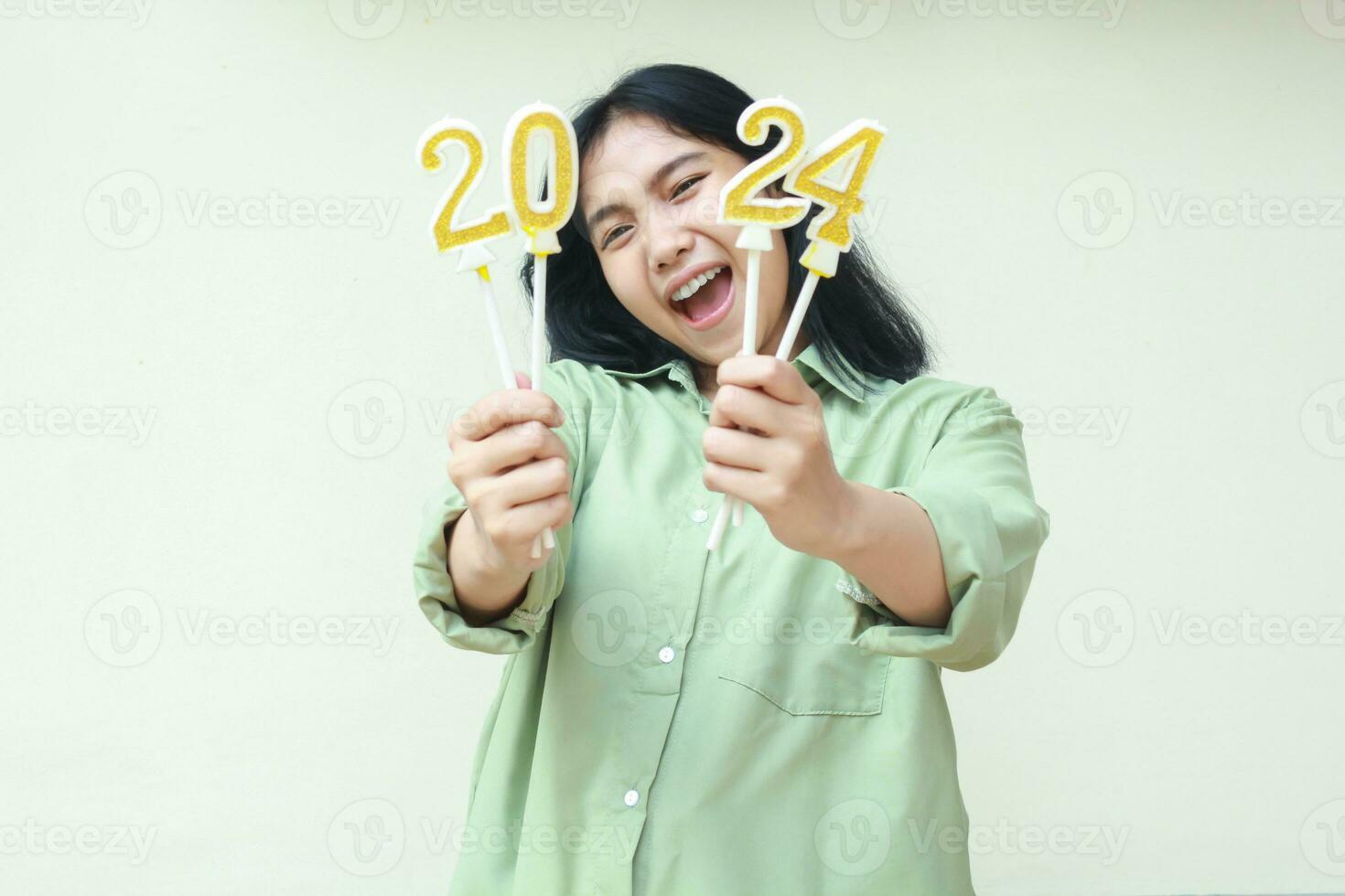 glücklich asiatisch jung Frau Hipster tragen Grün Über Größe Hemd schreiend zu Kamera mit erziehen 2024 Nummer Kerzen halten auf Hand, isoliert Über Weiß Hintergrund foto