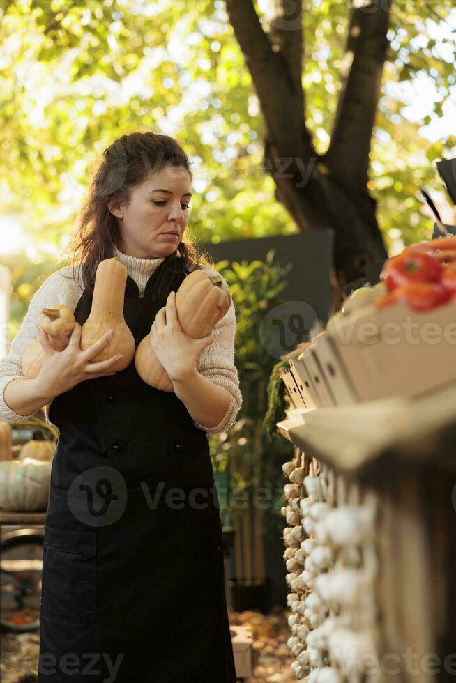 jung weiblich klein Geschäft Inhaber tragen schwarz Schürze mit Kürbisse im Hände Stehen in der Nähe von frisch organisch produzieren Stand beim Bauernhof Markt. weiblich Farmer Verkauf örtlich gewachsen Essen foto