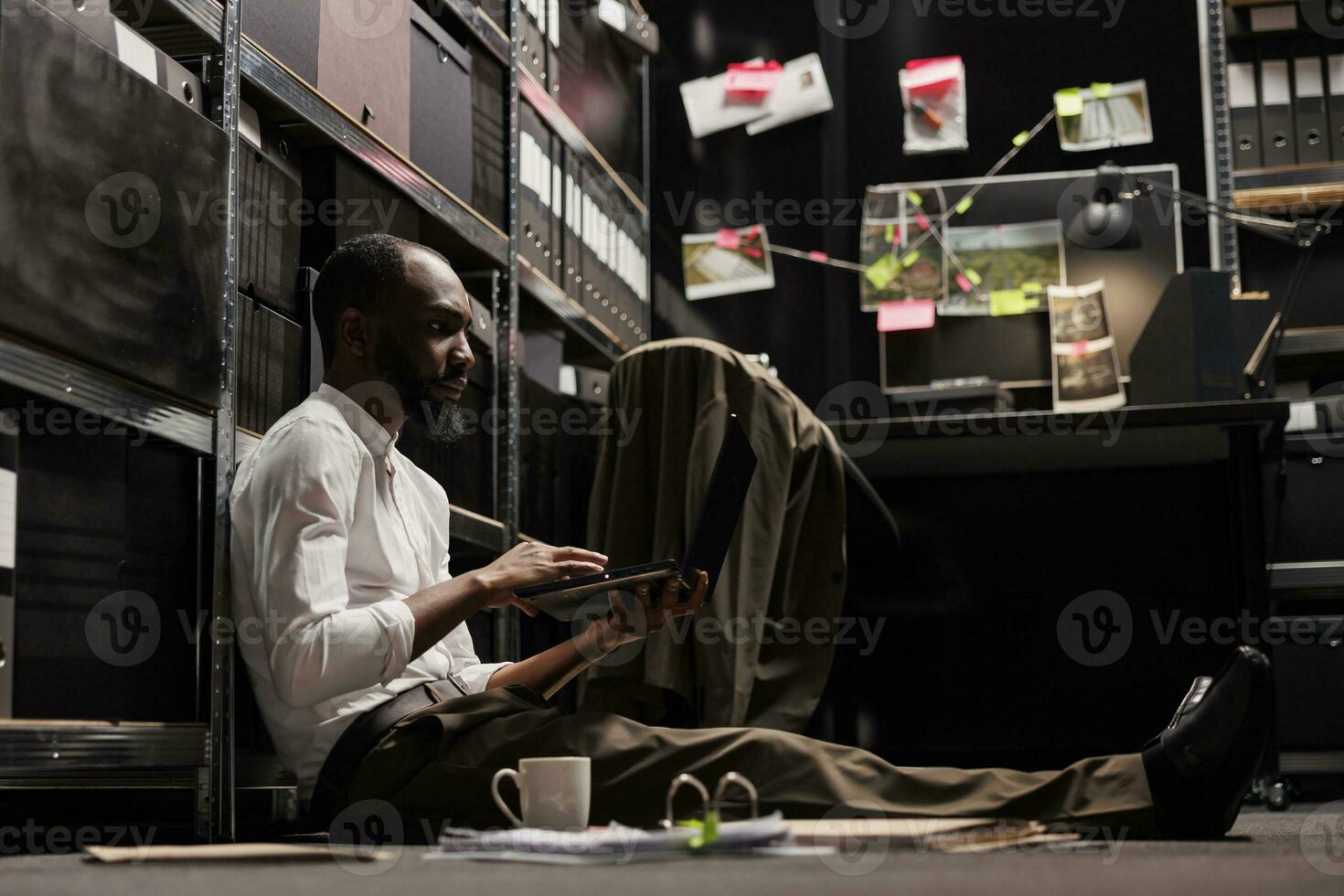afrikanisch amerikanisch Polizist Sitzung auf Fußboden und Arbeiten auf Laptop im Büro beim Nacht Zeit. Polizei Ermittler Prüfung Hinweise und vermuten Fotos auf Computer zu lösen Verbrechen Fall