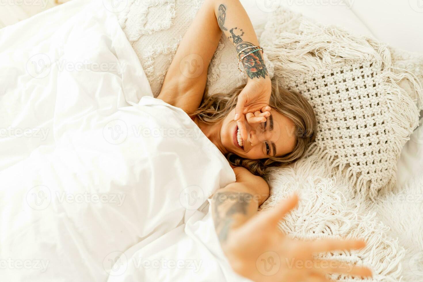 oben Aussicht Porträt von glücklich heiter Frau Lügen im Weiß gemütlich Bett aufwachen oben im das Morgen, zieht Hände nach vorne. warm Farben, Boho Innere. foto