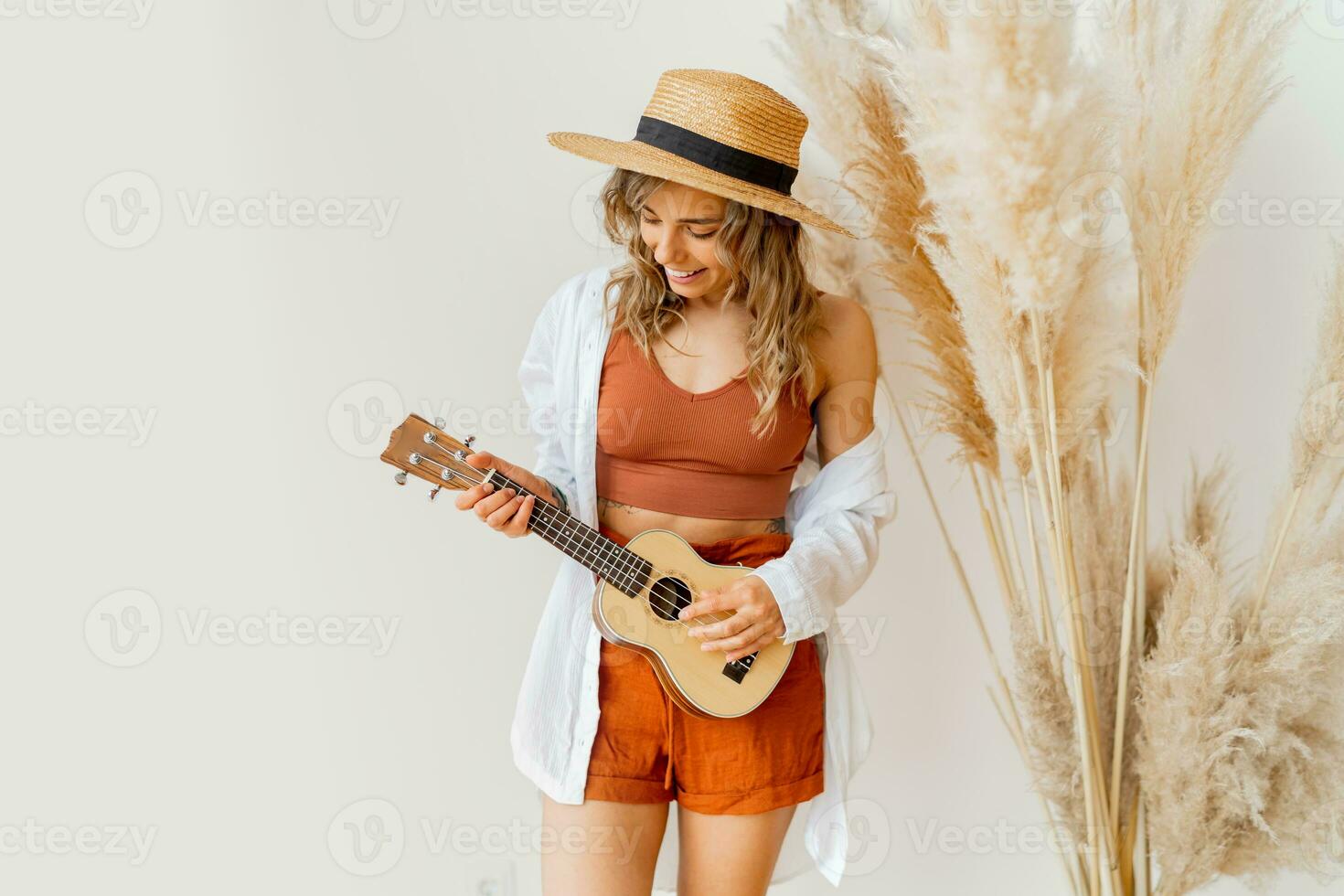 Innen- Foto von blond anmutig Frau im Sommer- Outfit mit Stroh Hut spielen Ukulele Gitarre Über Weiß Hintergrund im Studio mit Pampas Gras Dekor.