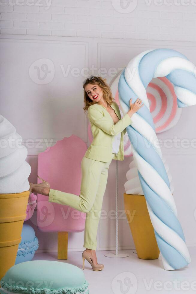 stilvoll elegant blond Frau posieren im Studio mit Süßigkeiten im beiläufig Pastell- passen . Süßigkeiten und Makronen Objekte Hintergrund. Sanft Pastell- Farben. foto