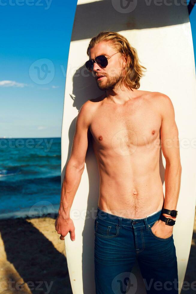 jung attraktiv Surfer Sport Mann mit lange Haar tragen Jeans kurze Hose und suchen beim das Horizont während Tragen seine Surfen Tafel während ein sonnig Tag gegen das heftig Blau Himmel. foto