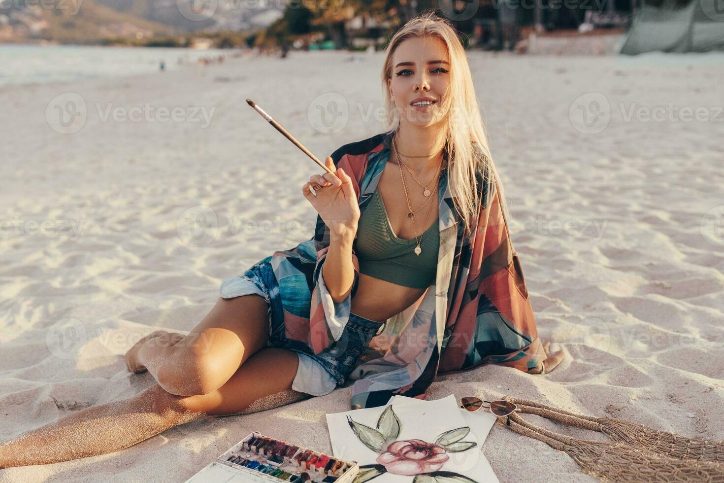 Sommer- Lebensstil Bild von blond Frau Zeichnung Aquarell Blume durch Bürste. Künstler Sitzung auf das Strand. Bohemien Outfit. foto