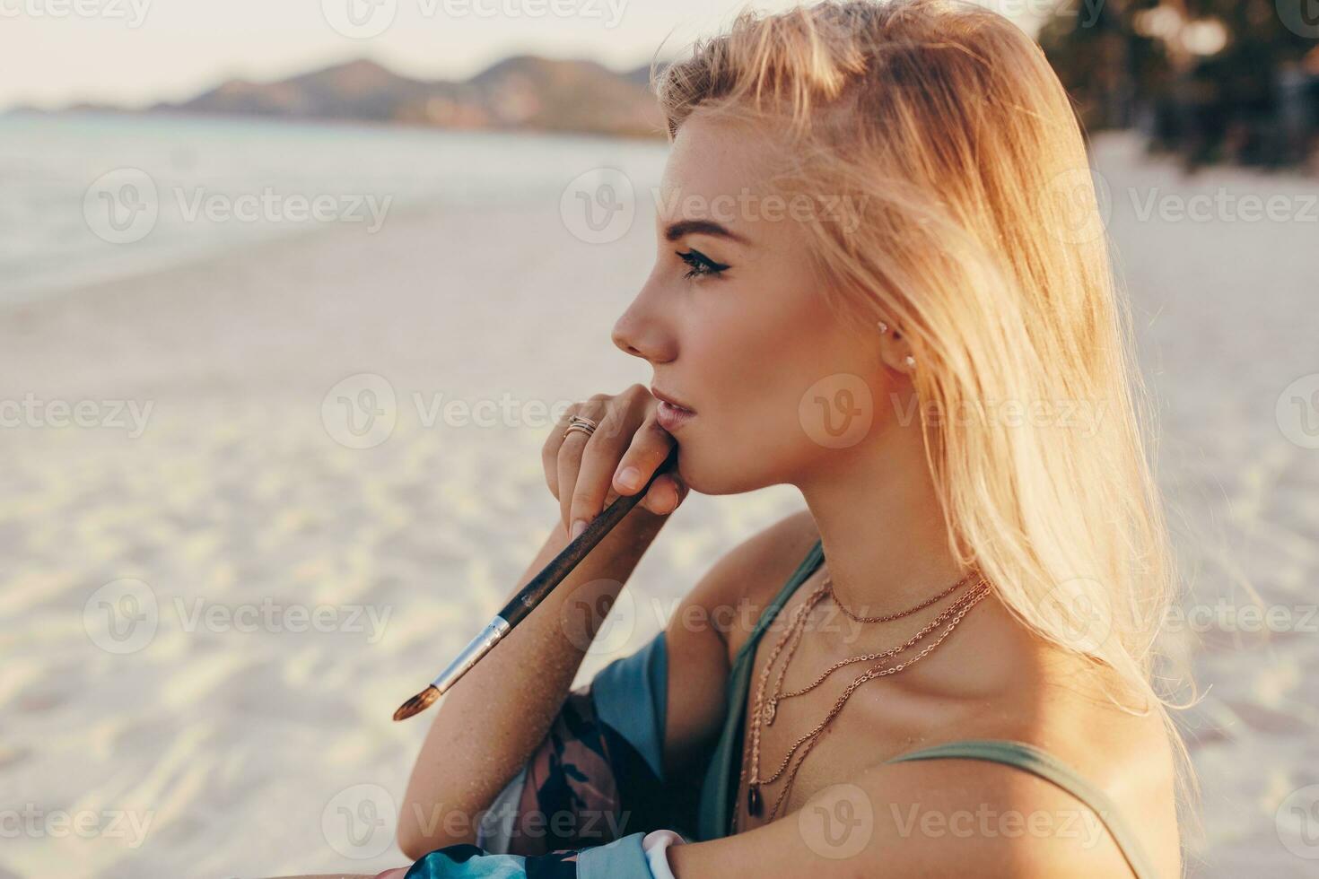 Sommer- Lebensstil Bild von blond Frau Zeichnung Aquarell Blume durch Bürste. Künstler Sitzung auf das Strand. Bohemien Outfit. foto