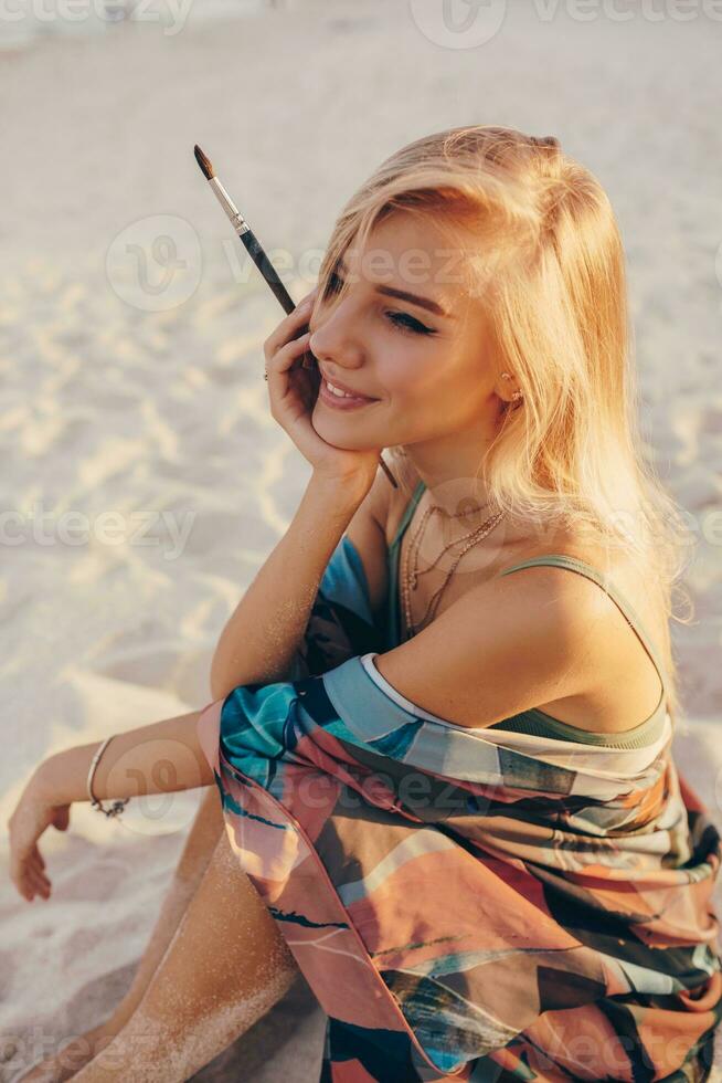 Sommer- Lebensstil Bild von blond Frau Zeichnung Aquarell Blume durch Bürste. Künstler Sitzung auf das Strand. Bohemien Outfit. foto