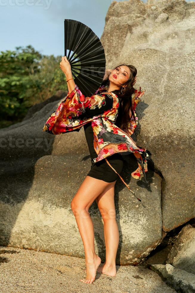 schön japanisch Frau im stilvoll Kimono mit groß Ventilator und Fachmann machen oben posieren Über Felsen. voll Länge. foto