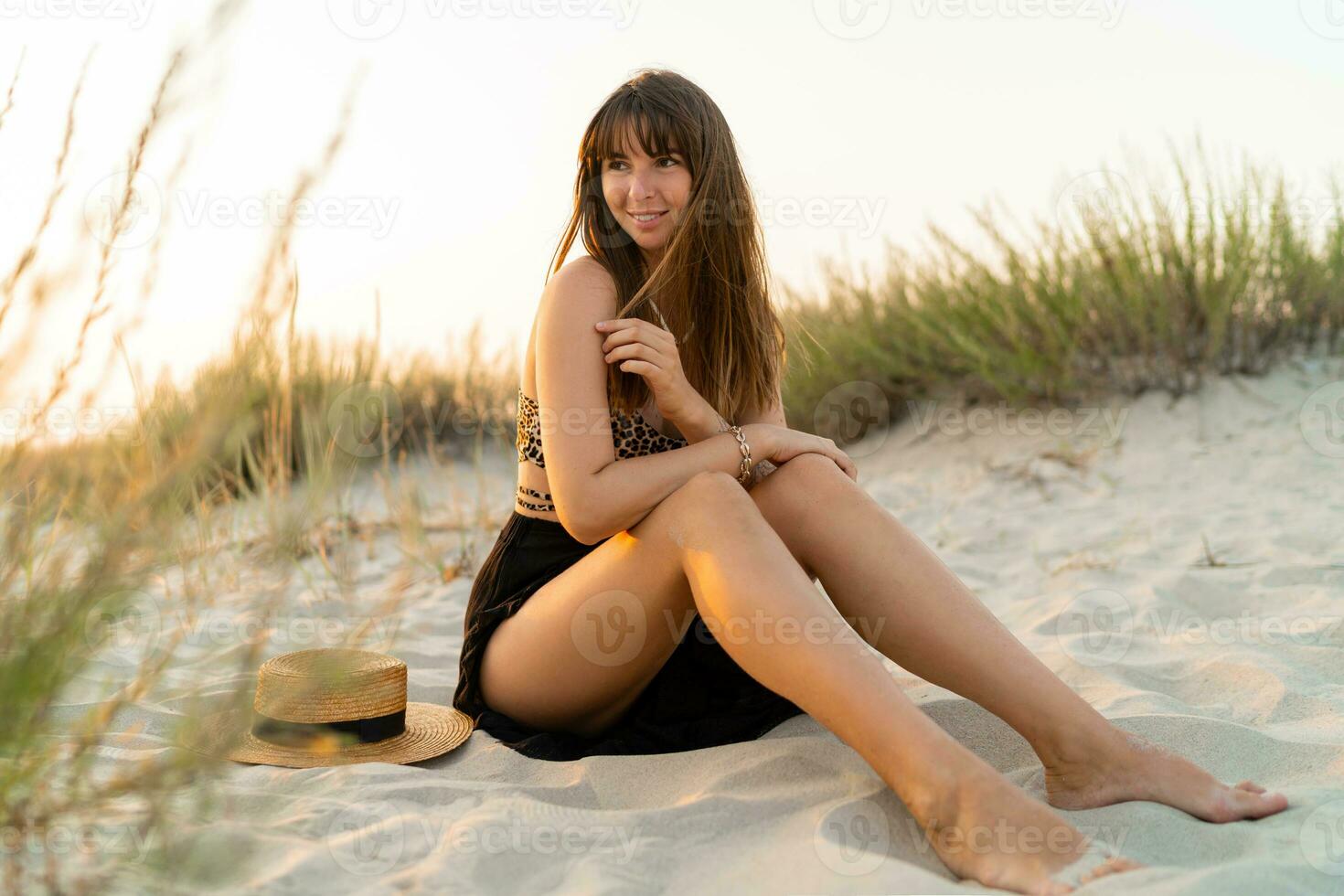 sexy Frau im stilvoll Sommer- Boho Outfit Sitzung auf Sand und genießen Ferien auf das Strand. Sonnenuntergang Farben. foto