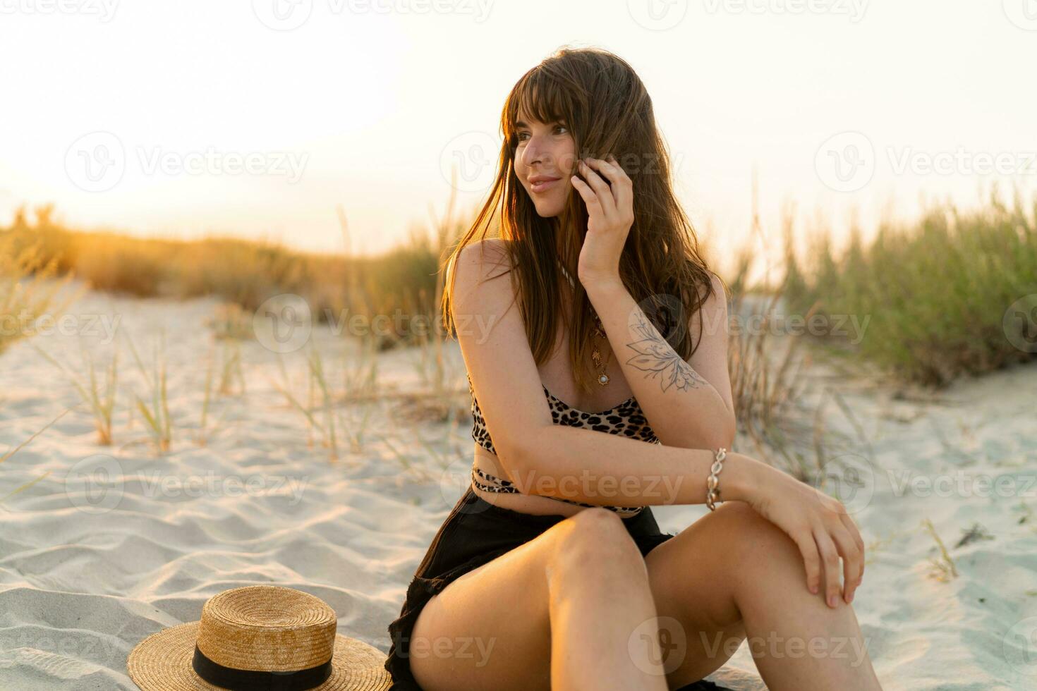 sexy Frau im stilvoll Sommer- Boho Outfit Sitzung auf Sand und genießen Ferien auf das Strand. Sonnenuntergang Farben. foto