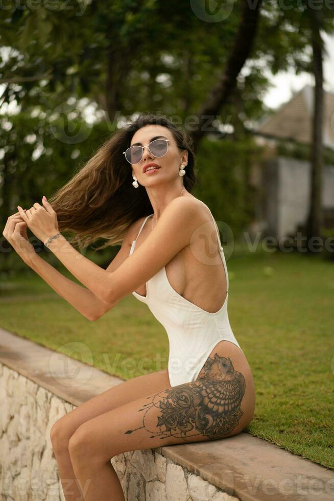 draussen Sommer- Mode Foto von elegant verführerisch Frau mit wellig Haare posieren auf tropisch Strand während Sommer- Urlaube. tragen Weiß Stilis Körper Bademode.