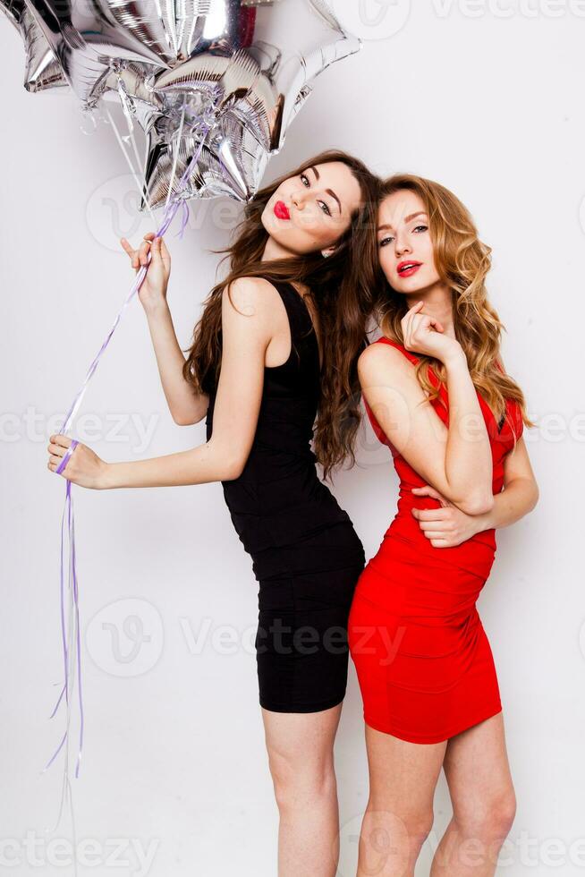 zwei schön elegant Frauen mit rot Lippen im Abend schwarz und rot Kleid haben Spaß. einer behalten rot Sterne Luftballons im ihr Hand und lächelnd. zwei Frau beim das Party. innen. Weiß Hintergrund. foto