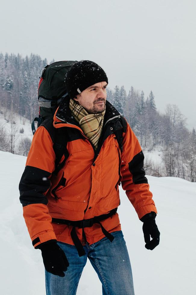touristischer Mann, der am Wintertag durch den Schnee geht, Bergwaldlandschaft. blaue Jeans, orangefarbenes Kleidungsstück, roter Rucksack. Wandern Reisen extremes Konzept. selektiver Fokus foto